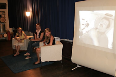 Theateraufführung "Fernsehgeschichten" mit Film auf einer Leinwand und Gesprächsgruppe in Sesseln