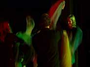 Szene aus der Revue. TnzerInnen schwingen Tcher auf einer in farbiges Licht getauchten Bhne