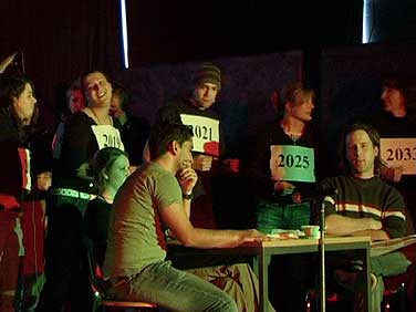 Szene aus der Revue. Chor mit Nummern hinter einem Tisch, an dem die Schauspieler einer gespielten Wohngemeinschaft sitzen.
