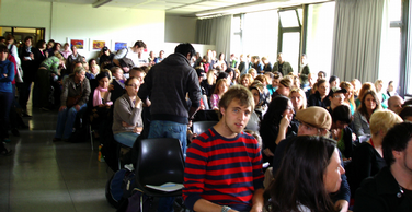Studierende sitzen im VV-Raum auf Stühlen und schauen nach vorne in die Richtung der Kamera.