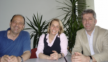 3 Personen sitzen an einem Tisch und lächeln fröhlich in die Kamera. Links sitzt der neue Dekan Prof Dr. Hubert Minkenberg in einem blauen Polo-Shirt. In der Mitte Prof. Dr. Charlotte Hanisch (Prodekanin) in einer weissen Bluse mit schwarzer Weste. Rechts sitzt Prof. Dr. Walter Eberlei (Prodekan) in einem blau-weiss gestreiftem Hemd und trägt dazu ein sandfarbenes Jacket.