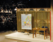 Auf der Bühne im FFT steht eine nachgebaute Kulisse einer Tagesschauaufnahme: ein Tisch mit Stuhl, links daneben eine Weltkarte und eine Kamera, die auf den Tisch gerichtet ist. Im Hintergrund kann man einige Publikumsgäste erkennen.