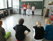 Das Bild zeigt eine drei Teilnehmende eines Theaterworkshops, die vor einer Gruppe etwas präsentieren.