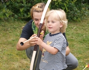 Auf diesem Bild sieht man ein Kind, welches einen Bogen hält. Eine Frau steht hinter ihm und hilft ihm, den Bogen zu halten.