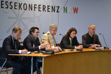 Mehrere Personen sitzen auf dem Podium an einem halbrunden Tisch, hinter Ihnen ist eine Wand mit der Aufschrift "Landespressekonferenz NRW"