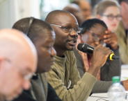 Das Bild zeigt einen Teilnehmer mit Mikrofon in der Hand, der sich an einer plenaren Diskussion im Rahmen der Winter School beteiligt. Er sitzt zwischen mehreren anderen Teilnehmer_innen an einem Tisch.