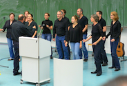 Der Pop- und Jazzchor der FHD singt mit elf Personen mit dunklen Hemden bekleidet vor der Tafel im Hörsaal unter der Leitung von Prof. Dr. H. Minkenberg bei der Abschlussfeier des FB6.