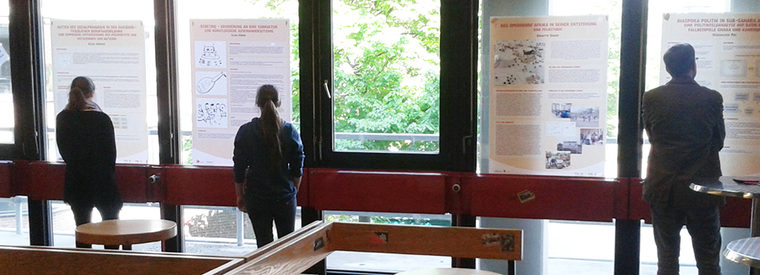 Das Bild zeigt in Form eines Panoramas die im Artikel beschriebene Ausstellung im Foyer, während diese von Studierenden betrachtet wird.