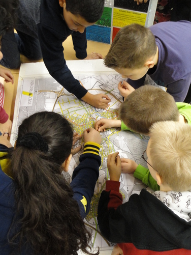 Das Foto zeigt Kinder, die über einen großen auf dem Boden liegenden Stadtplan gebeugt bestimmte Stellen darin markieren.