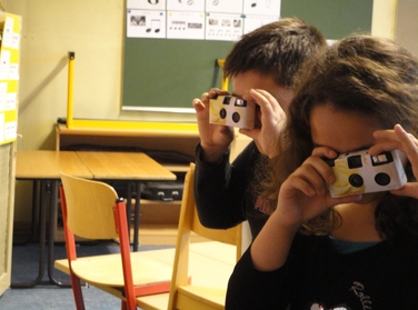 Zwei Kinder mit Einwegfotoapparaten vor dem Gesicht in einer Schulklasse