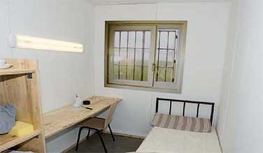 Gefngnisszelle mit Pritsche, Holztisch und vergittertem Fenster
